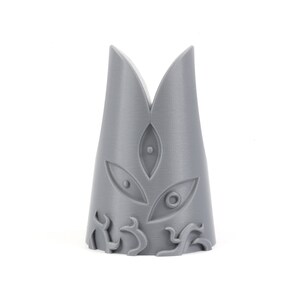 May include: A gray 3D printed crown with a pointed top and three eye-like designs on the front. The crown has a decorative design at the base.