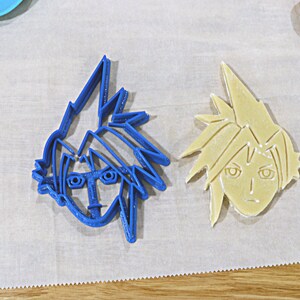 May include: Two cookie cutters, one blue and one beige, shaped like the face of a cartoon character with spiky hair.