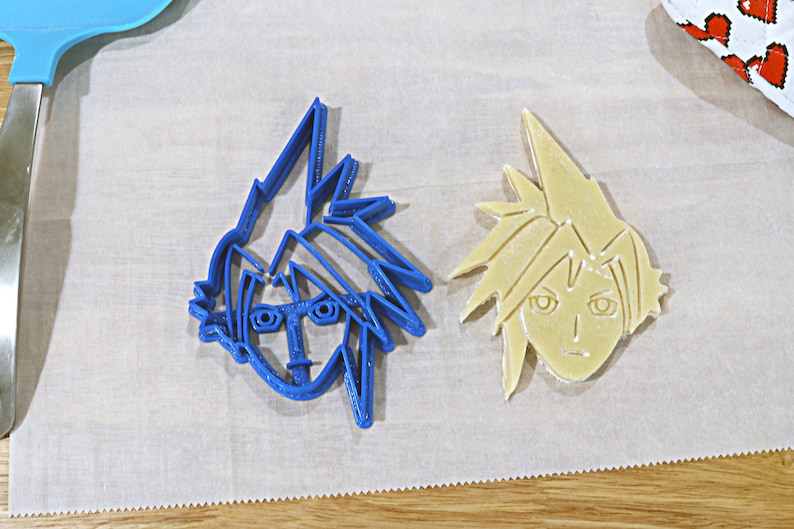 May include: Two cookie cutters, one blue and one beige, shaped like the face of a cartoon character with spiky hair.