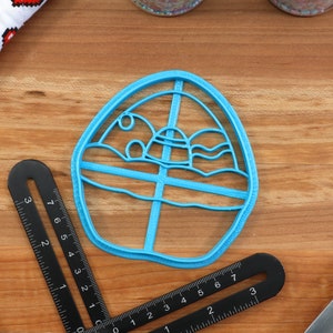 May include: A blue plastic cookie cutter shaped like a cartoon egg with a sun, clouds, and waves inside. The cutter is on a wooden surface.