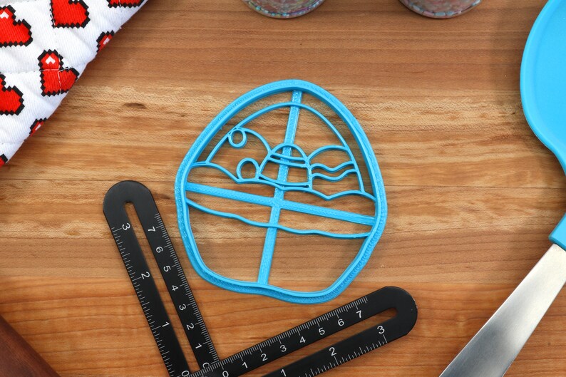 May include: A blue plastic cookie cutter shaped like a cartoon egg with a sun, clouds, and waves inside. The cutter is on a wooden surface.