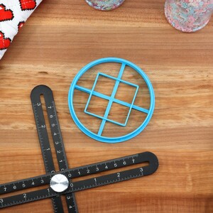 May include: A top-down shot of baking tools on a wooden surface. A blue plastic cookie cutter with a square design sits in the center. A black multi-angle measuring tool is below it. A white and red heart-patterned cloth is in the upper left.