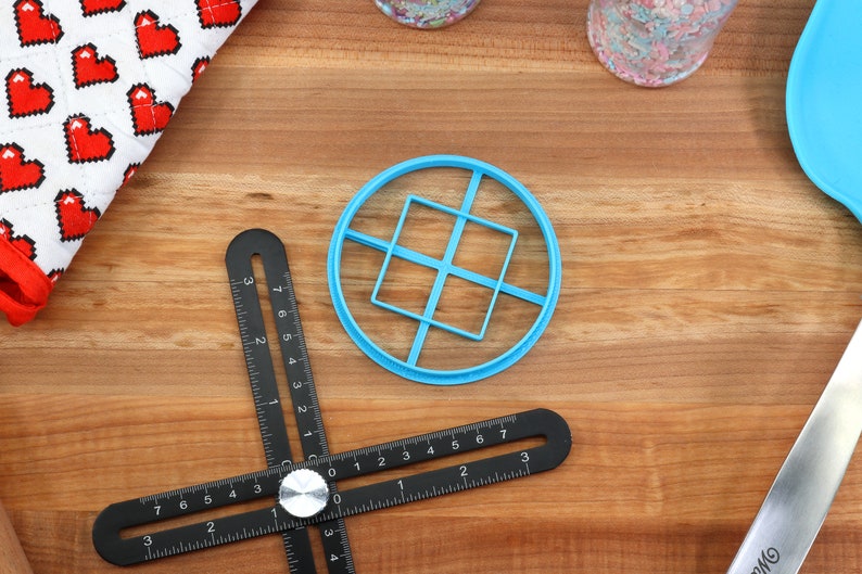 May include: A top-down shot of baking tools on a wooden surface. A blue plastic cookie cutter with a square design sits in the center. A black multi-angle measuring tool is below it. A white and red heart-patterned cloth is in the upper left.