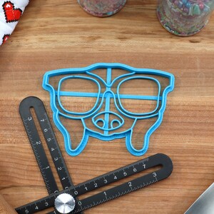 May include: Blue plastic cookie cutter shaped like a dog wearing glasses. The cookie cutter is on a wooden surface.