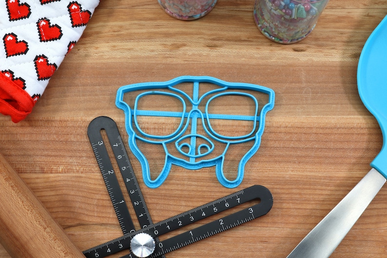 May include: Blue plastic cookie cutter shaped like a dog wearing glasses. The cookie cutter is on a wooden surface.