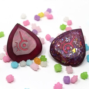 May include: Two teardrop-shaped, burgundy-colored pendants with a pink symbol are displayed. One is solid, the other glittery. They are surrounded by colorful, star-shaped candies on a white surface. The pendants are likely jewelry.