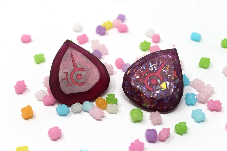 May include: Two teardrop-shaped, burgundy-colored pendants with a pink symbol are displayed. One is solid, the other glittery. They are surrounded by colorful, star-shaped candies on a white surface. The pendants are likely jewelry.