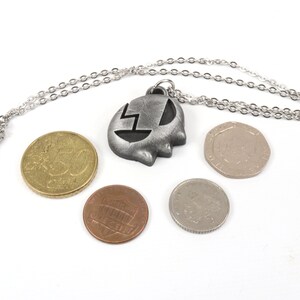 May include: A silver necklace with a pendant shaped like a ghost with a lightning bolt inside. The necklace is laying on a white surface with four coins.
