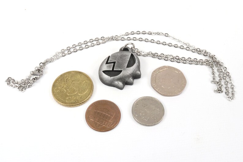 May include: A silver necklace with a pendant shaped like a ghost with a lightning bolt inside. The necklace is laying on a white surface with four coins.