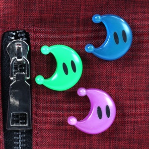 May include: Three crescent moon-shaped plastic toys with black eyes. The toys are blue, green, and purple and are arranged on a red fabric surface.