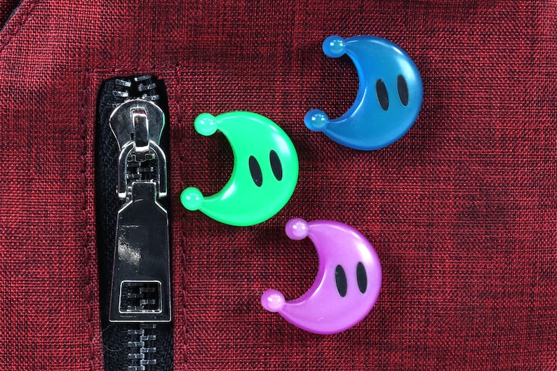 May include: Three crescent moon-shaped plastic toys with black eyes. The toys are blue, green, and purple and are arranged on a red fabric surface.