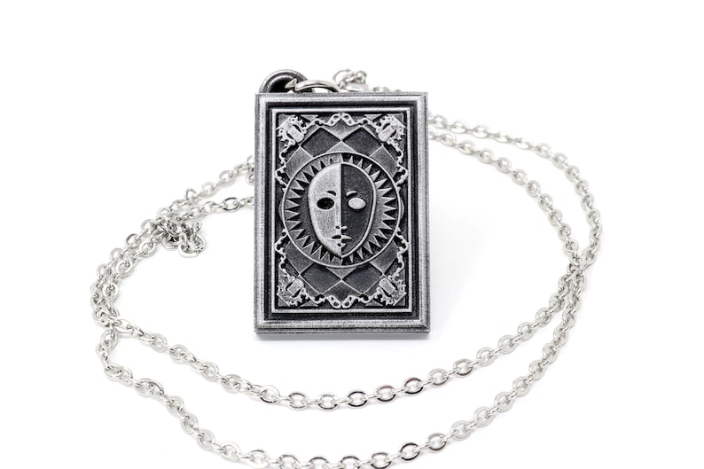 Puede incluir: Un collar de plata con un colgante que presenta un dise&ntilde;o de m&aacute;scara facial en blanco y negro. El colgante es rectangular y tiene un borde detallado. La cadena es de plata y tiene un eslab&oacute;n delicado.