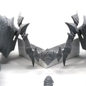 May include: A pair of gray 3D printed dragon head sculptures with sharp teeth and horns. The sculptures are facing each other and are positioned on a white surface.
