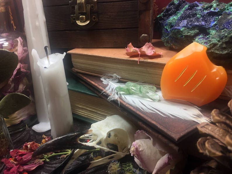 May include: A still life featuring a white candle, a stack of old books, a white feather, a green crystal, an orange carved stone, and a dried flower.