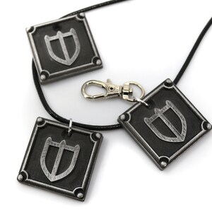 May include: Three silver-coloured pendants with a shield design on a black cord necklace. The pendants are square-shaped with rounded corners and have a silver-coloured metal clasp.