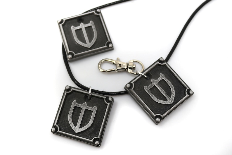 May include: Three silver-coloured pendants with a shield design on a black cord necklace. The pendants are square-shaped with rounded corners and have a silver-coloured metal clasp.