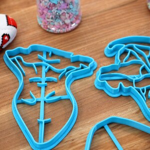 May include: Three blue plastic cookie cutters on a wooden surface. The cutters are shaped like a heart, a flower, and a rectangle.