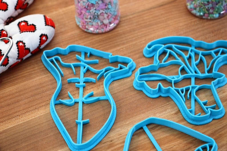 May include: Three blue plastic cookie cutters on a wooden surface. The cutters are shaped like a heart, a flower, and a rectangle.