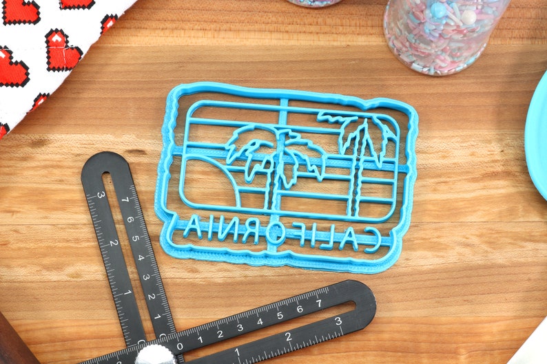 May include: Blue plastic cookie cutter shaped like a California state outline with palm trees and the word "CALIFORNIA" written inside.