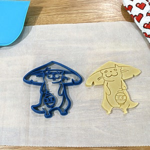 May include: Two cookie cutters, one blue and one yellow, shaped like a cartoon cat wearing a hat and holding a lantern. The cutters are on a white parchment paper.