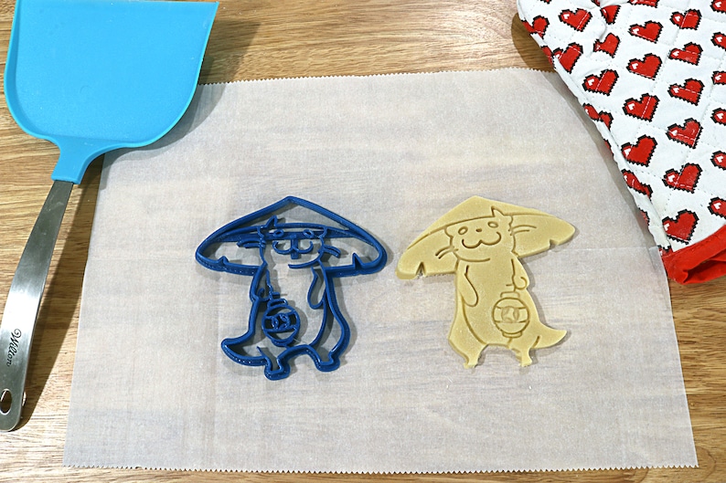 May include: Two cookie cutters, one blue and one yellow, shaped like a cartoon cat wearing a hat and holding a lantern. The cutters are on a white parchment paper.