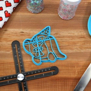 May include: A blue plastic cookie cutter in the shape of a cartoon dog wearing a party hat, with a bow tie. Also visible are a blue spatula, jars of sprinkles, a heart-patterned oven mitt, and a black measuring tool, all on a wooden surface.