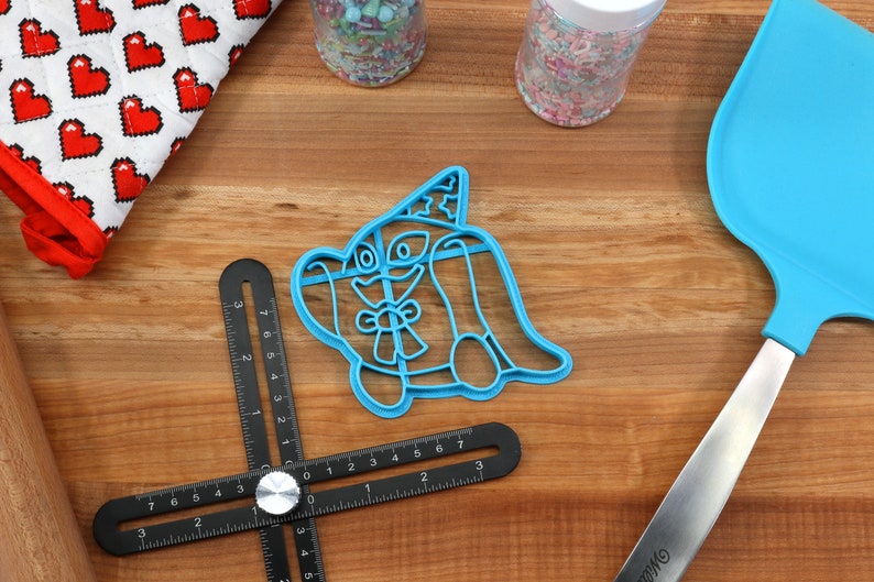 May include: A blue plastic cookie cutter in the shape of a cartoon dog wearing a party hat, with a bow tie. Also visible are a blue spatula, jars of sprinkles, a heart-patterned oven mitt, and a black measuring tool, all on a wooden surface.