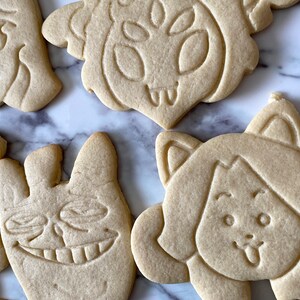 May include: Five sugar cookies decorated with various cartoon faces. The cookies are light brown and have a slightly rough texture.