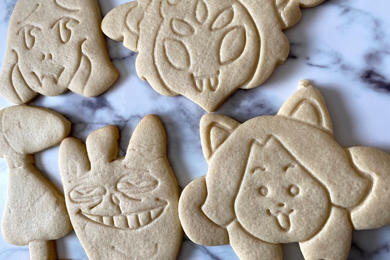 May include: Five sugar cookies decorated with various cartoon faces. The cookies are light brown and have a slightly rough texture.