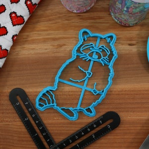 Puede incluir: Un cortador de galletas de pl&aacute;stico azul con forma de mapache. El cortador de galletas est&aacute; sobre una superficie de madera.