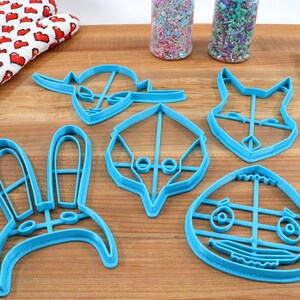 May include: Five blue plastic cookie cutters shaped like animal faces. The cutters are on a wooden surface.