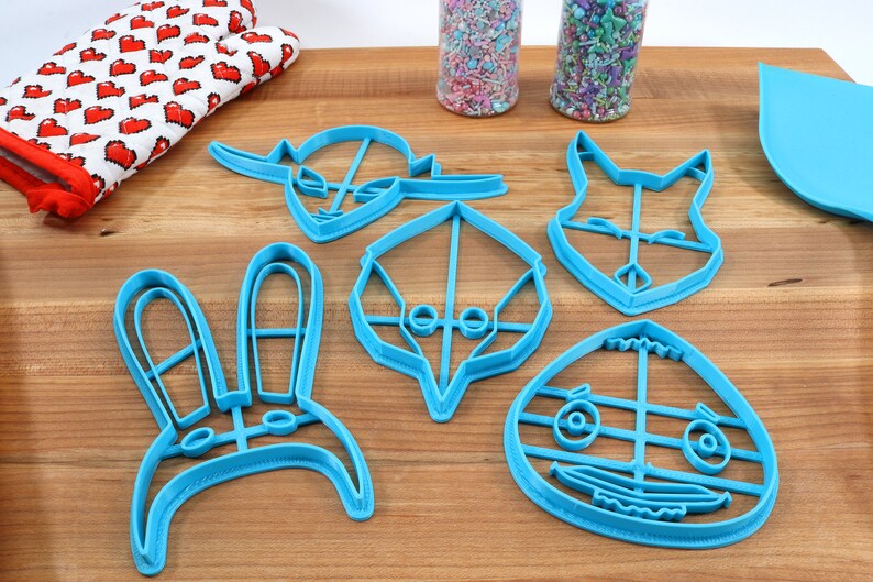 May include: Five blue plastic cookie cutters shaped like animal faces. The cutters are on a wooden surface.