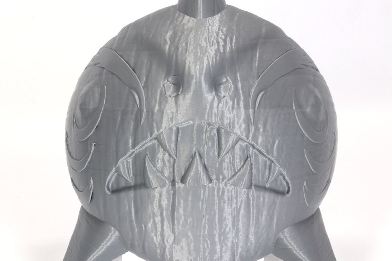 May include: A gray 3D printed sculpture of a creature with a toothy grin and large eyes. The sculpture is detailed with textured lines and a textured surface.