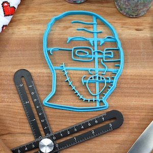 May include: A blue plastic cookie cutter shaped like a person's face with glasses and a beard. The cookie cutter is on a wooden surface.