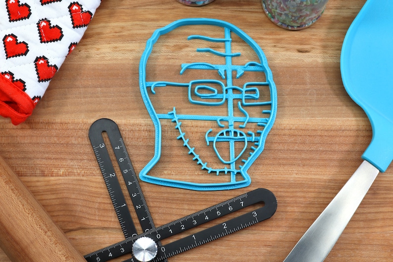 May include: A blue plastic cookie cutter shaped like a person's face with glasses and a beard. The cookie cutter is on a wooden surface.