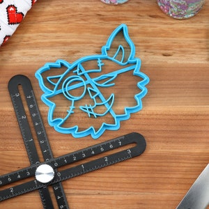 May include: A blue plastic cookie cutter shaped like a cartoon monster face with stitches and a single eye. The cutter is on a wooden surface with a black ruler and a blue spatula.