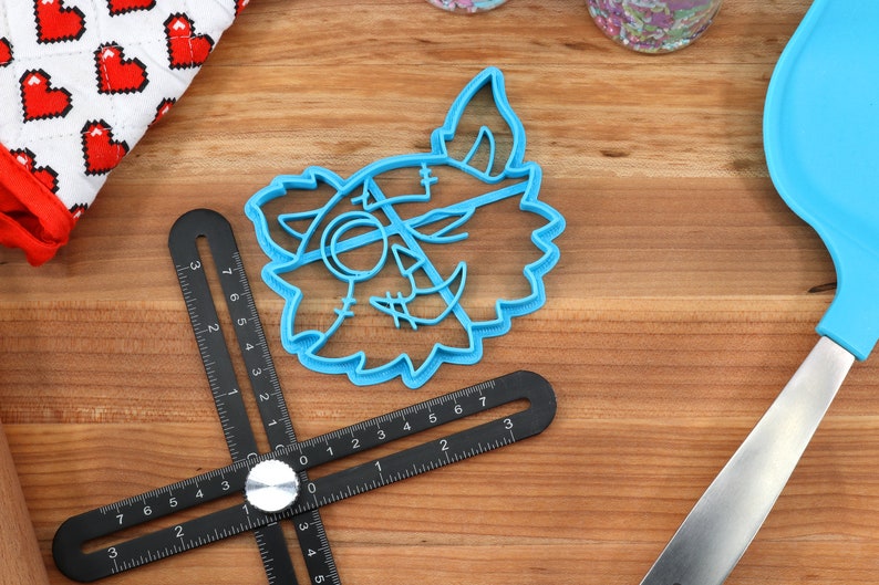 May include: A blue plastic cookie cutter shaped like a cartoon monster face with stitches and a single eye. The cutter is on a wooden surface with a black ruler and a blue spatula.