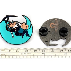 May include: A teal blue enamel pin featuring an anime-style character holding a scythe, sitting on a crescent moon. The pin is attached to a black metal backing with two butterfly clasps. The back of the pin has the text "LOOT CAVE CO. lootcave.com" in a brown font.