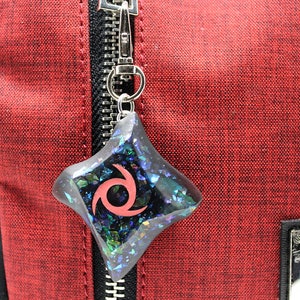 Pu&ograve; includere: Un ciondolo con un design a spirale nero e rosso con glitter iridescente incorporato in resina trasparente. Il ciondolo &egrave; attaccato a un portachiavi argentato e pende da una cerniera su una borsa rossa.