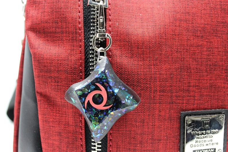 Pu&ograve; includere: Un ciondolo con un design a spirale nero e rosso con glitter iridescente incorporato in resina trasparente. Il ciondolo &egrave; attaccato a un portachiavi argentato e pende da una cerniera su una borsa rossa.