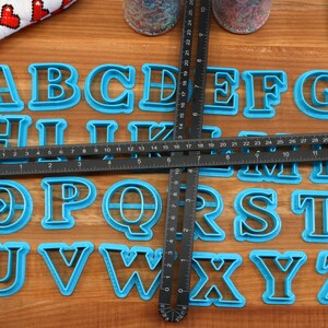 May include: A set of blue plastic alphabet cookie cutters on a wooden surface. The cutters are arranged in alphabetical order and are surrounded by a black ruler.