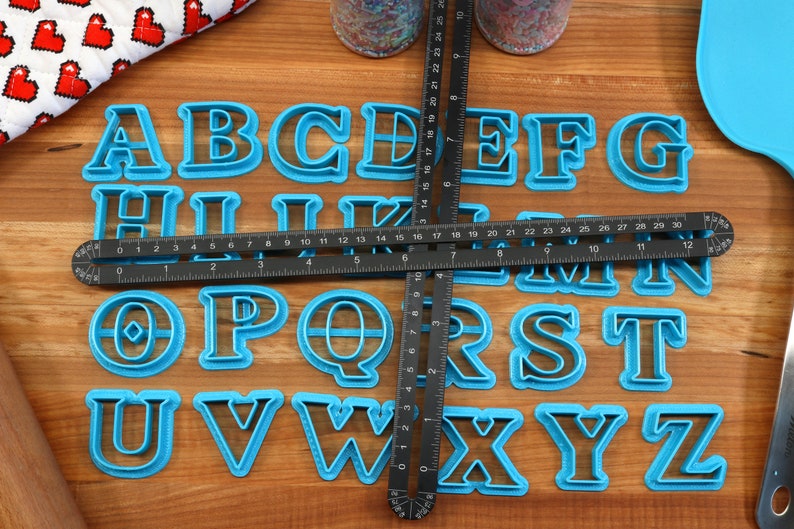 May include: A set of blue plastic alphabet cookie cutters on a wooden surface. The cutters are arranged in alphabetical order and are surrounded by a black ruler.