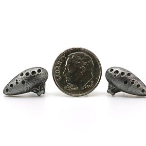 Pu&ograve; includere: Due piccoli ciondoli argentati a forma di ocarina con pi&ugrave; fori, che affiancano una moneta da dieci centesimi americana. I ciondoli hanno un aspetto invecchiato. La moneta mostra un profilo di uomo e la parola "Liberty".