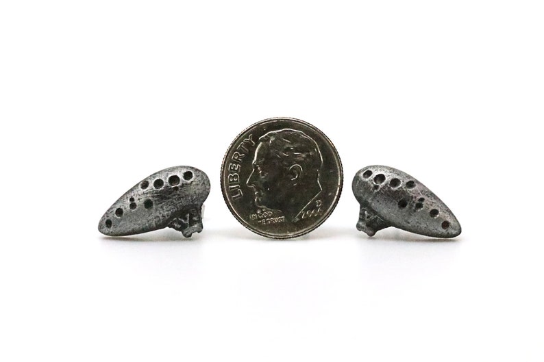Pu&ograve; includere: Due piccoli ciondoli argentati a forma di ocarina con pi&ugrave; fori, che affiancano una moneta da dieci centesimi americana. I ciondoli hanno un aspetto invecchiato. La moneta mostra un profilo di uomo e la parola "Liberty".