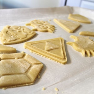 Puede incluir: Una bandeja para hornear con galletas de az&uacute;car sin hornear cortadas en varias formas, incluyendo una espada, un escudo, un tri&aacute;ngulo, un diamante y una estrella. Las galletas est&aacute;n sobre papel de pergamino blanco.