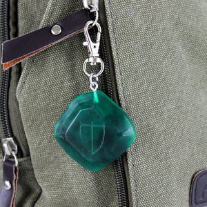 May include: A green, diamond-shaped keychain with a silver clasp and a shield design. The keychain is attached to a brown leather strap.