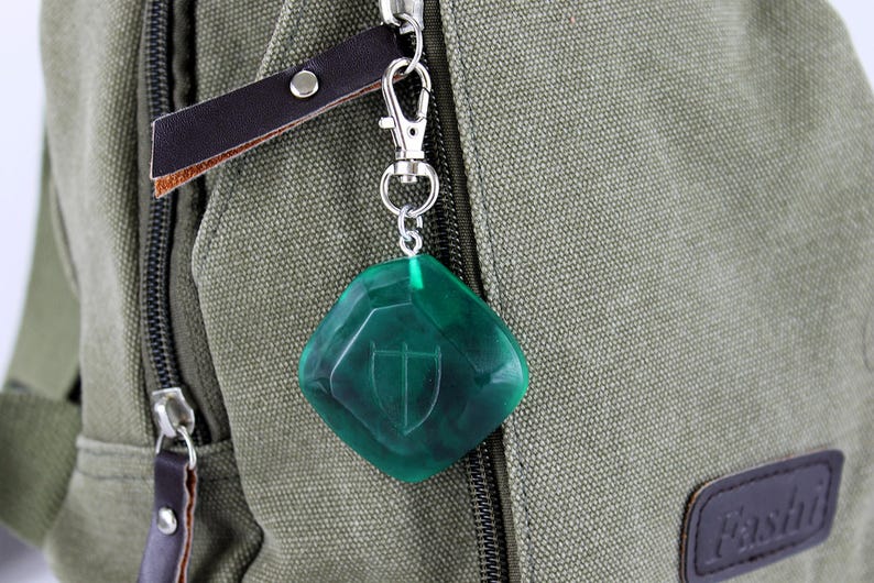 May include: A green, diamond-shaped keychain with a silver clasp and a shield design. The keychain is attached to a brown leather strap.