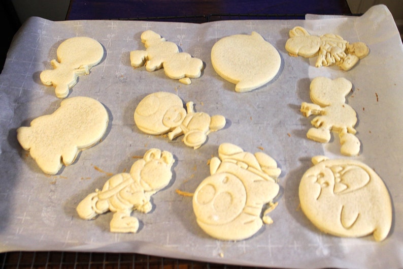 May include: A sheet of parchment paper with nine unbaked sugar cookies in various shapes, including a ghost, a character with a hat, and a character with a backpack.