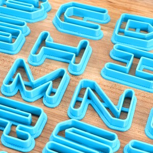 May include: A set of blue plastic cookie cutters shaped like letters of the alphabet. The cutters are arranged on a wooden surface.