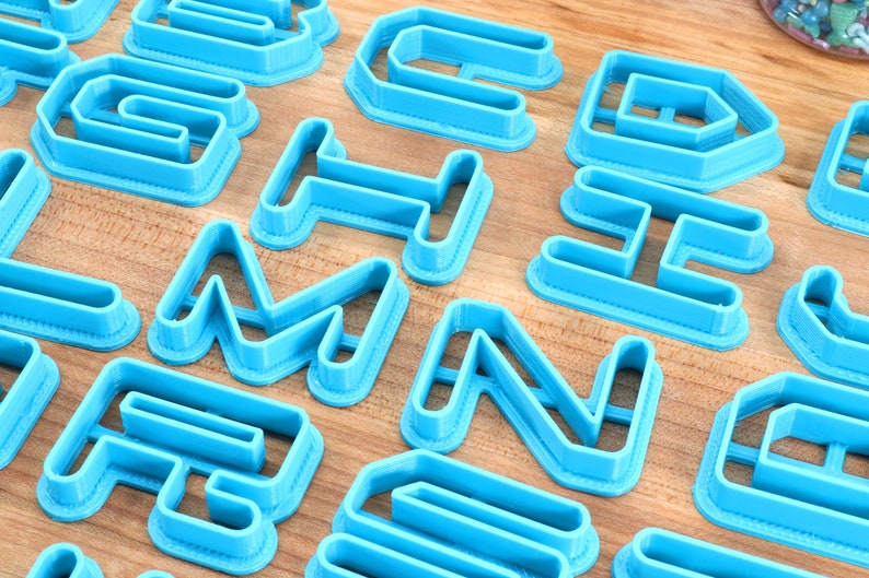 May include: A set of blue plastic cookie cutters shaped like letters of the alphabet. The cutters are arranged on a wooden surface.